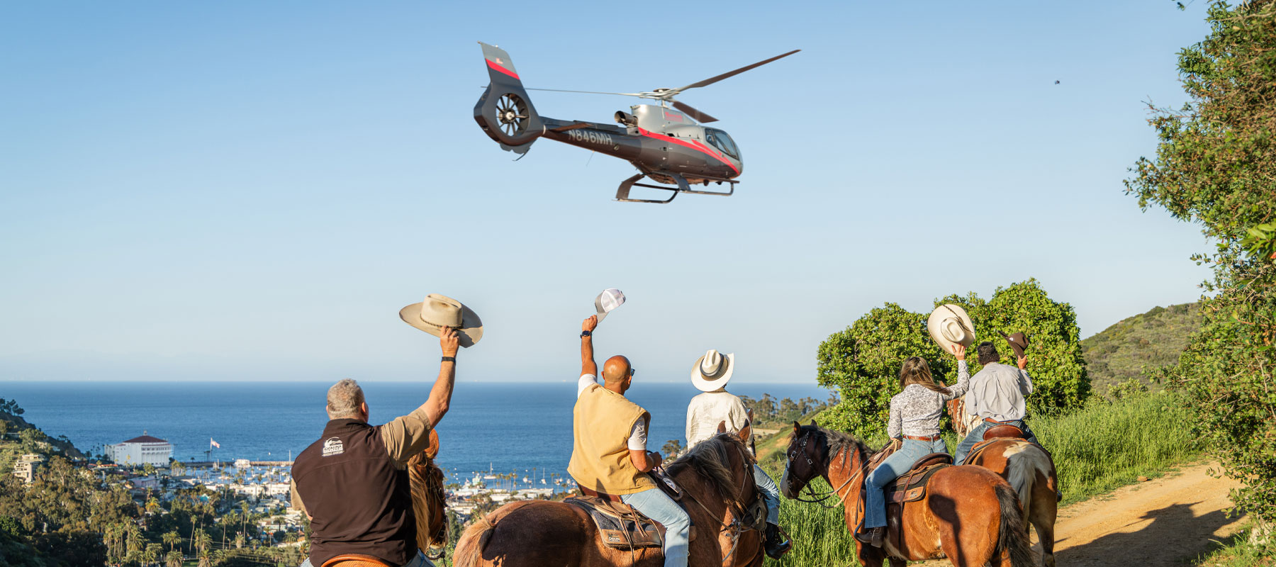 Riders on horseback waving at a helicopter flying over the Catalina Island coastline